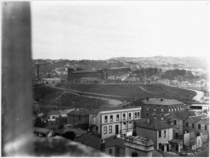 Alexandra Barracks, from the St Patrick’s College tower. The police station and associated stables are located to the right of the image. The open land behind the Tasman Street Wall is in use as horse paddocks. The barracks buildings can be seen at the centre of the image, and the chimney for the brickworks can be seen at the top-left.  National Library reference: Mount Cook Prison and buildings on Buckle Street, Wellington. Smith, Sydney Charles, 1888-1972 :Photographs of New Zealand. Ref: 1/1-020192-G. Alexander Turnbull Library, Wellington, New Zealand. /records/22410901